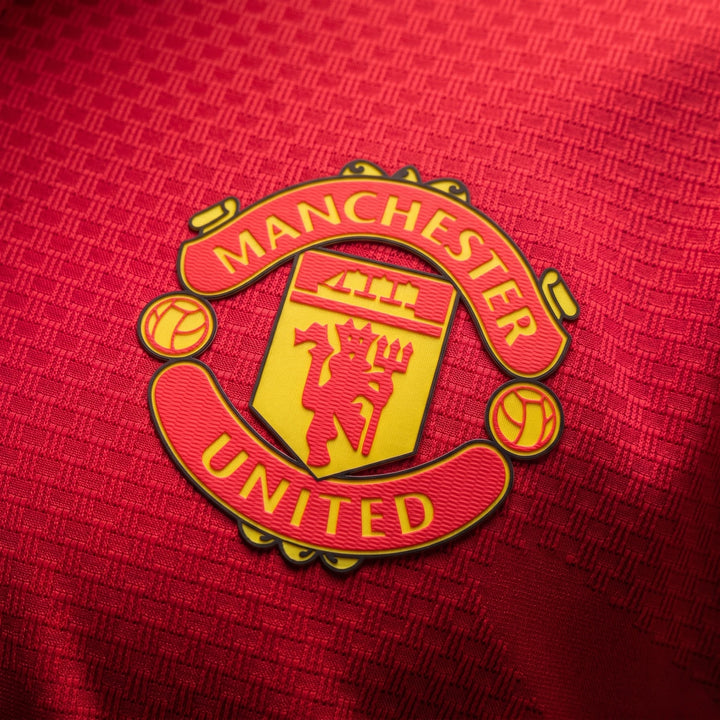 Manchester United 26/27 Authentic Long Sleeve Home Jersey, close-up of yellow crest badge on red textured fabric.