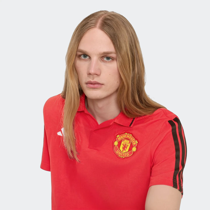 A male model with long blonde hair wears a red Manchester United Dna Polo Shirt with a club crest and black sleeve stripes.