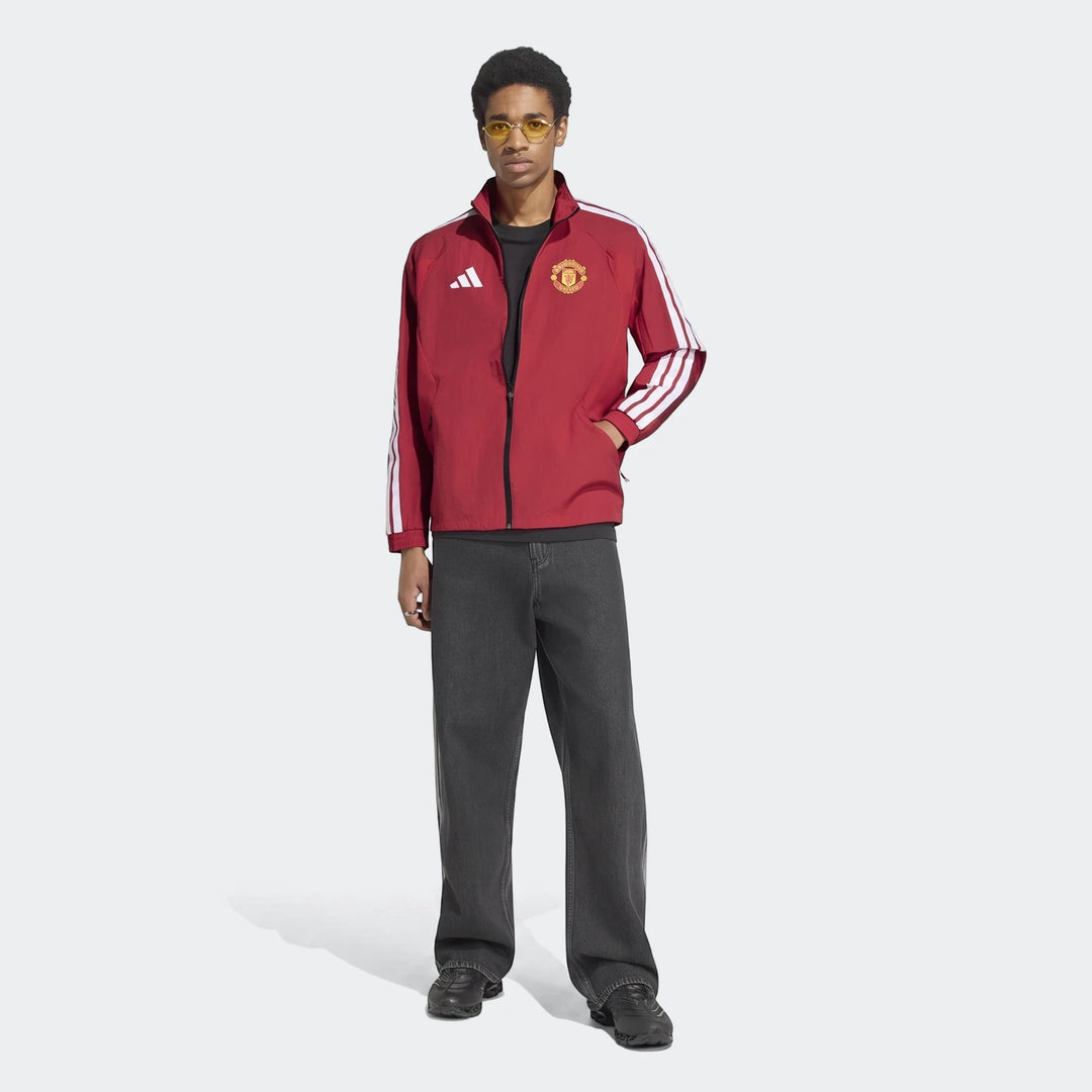 A man wears a red Manchester United Home Anthem Jacket featuring white Adidas stripes and a full-zip closure, styled with