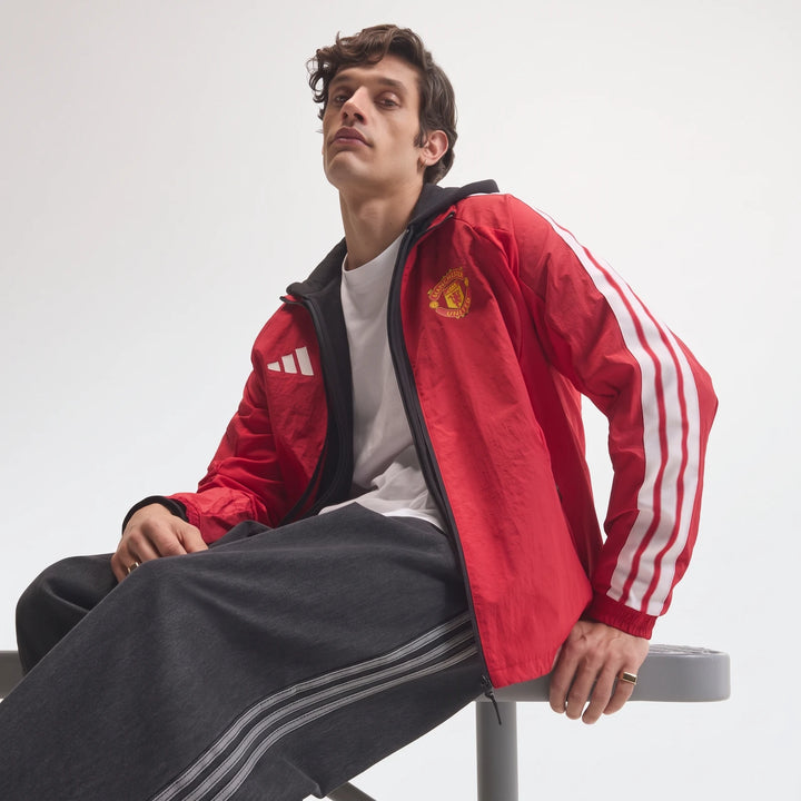 Male model wearing a red Manchester United Home Anthem Jacket with white 3-stripes and club crest, seated casually.