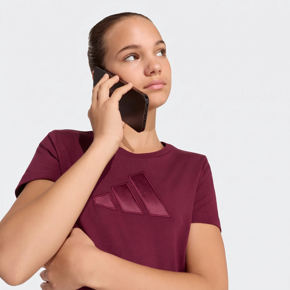 Young female model wearing a maroon Adidas Satin T-Shirt, featuring a prominent logo, holding a phone to her ear and looking