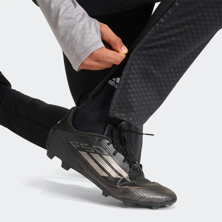 Men's black Adidas Tiro 26 Competition Training Pants with a zipper detail, worn with black soccer cleats, being adjusted by