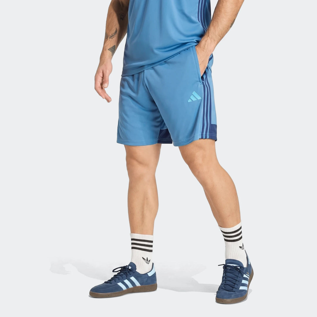 Man wearing blue Adidas Tiro Essentials Shorts, a blue t-shirt, and blue sneakers, captured from mid-torso to feet.