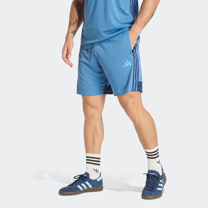 Man wearing blue Adidas Tiro Essentials Shorts, a blue t-shirt, and blue sneakers, captured from mid-torso to feet.