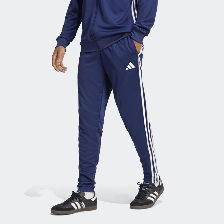 Men's blue Adidas Tiro Essentials track pants with white stripes worn with matching jacket and black sneakers.