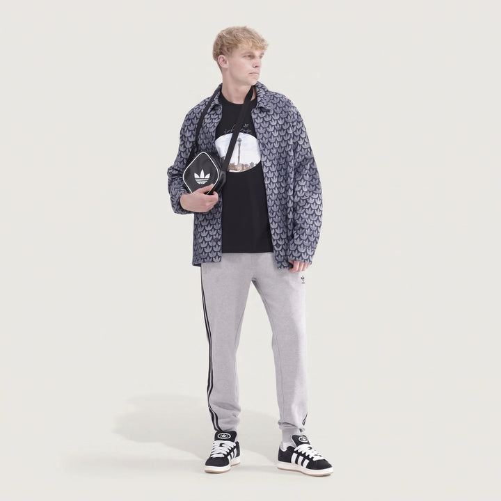Male model in full body shot wearing grey Adidas 3-Stripes Pants, a patterned jacket, and sneakers, holding a black Adidas