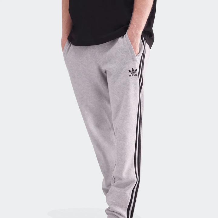 Men's grey Adidas 3-Stripes sweatpants, shown worn by a model with hands in pockets. Features black triple stripes and a
