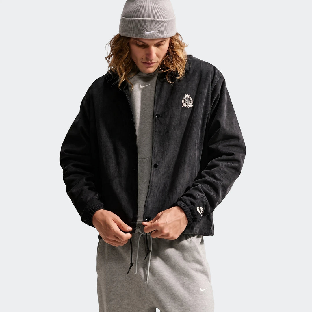 Men's Nike Club Corduroy Coaches Jacket in black, paired with a gray Nike hoodie, sweatpants, and beanie, worn by a male