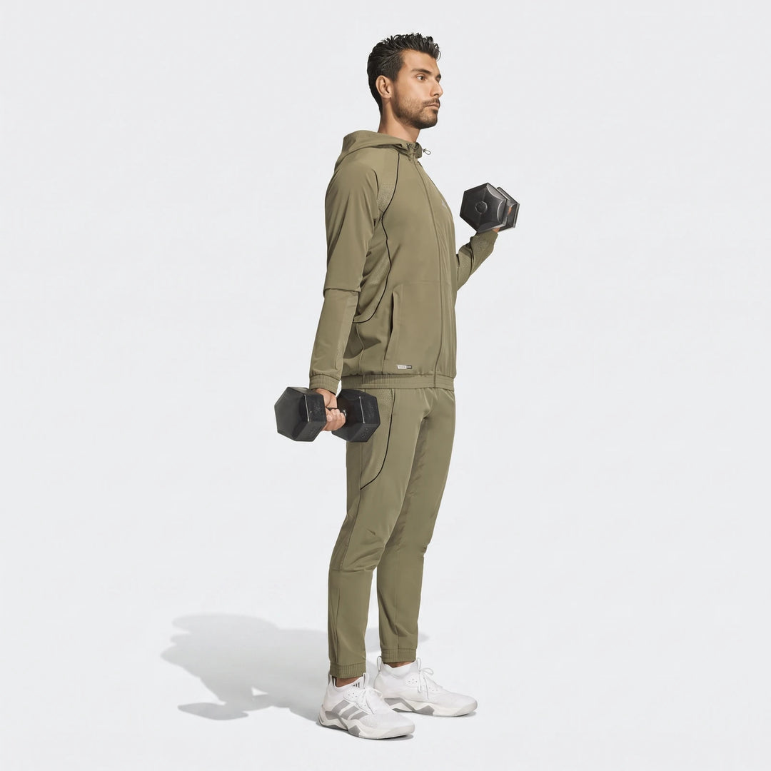 Full-body shot of a man wearing olive green technical apparel, including a hooded jacket and slim pants, holding dumbbells