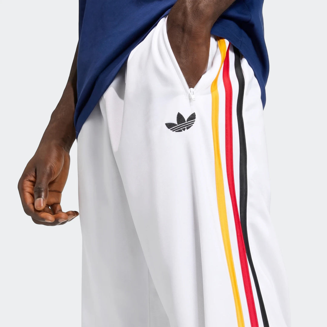 Close-up of men's white Adidas Firebird track pants with yellow, red, and black side stripes, showing a zippered pocket and