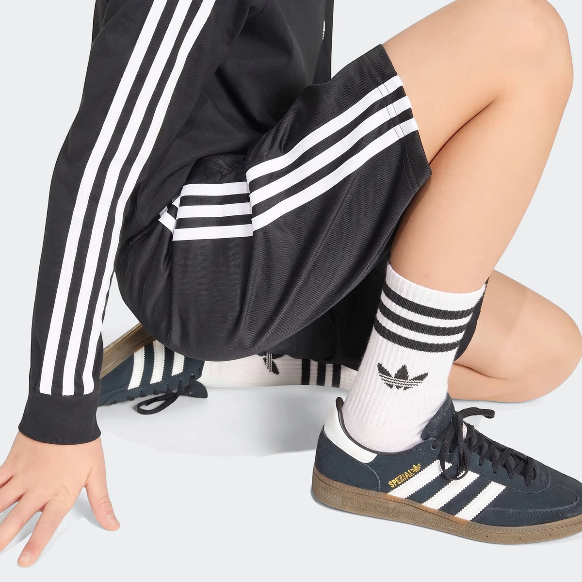 Model wearing black Firebird shorts with white stripes, a black long-sleeve shirt, and blue Adidas Spezial shoes, crouching.
