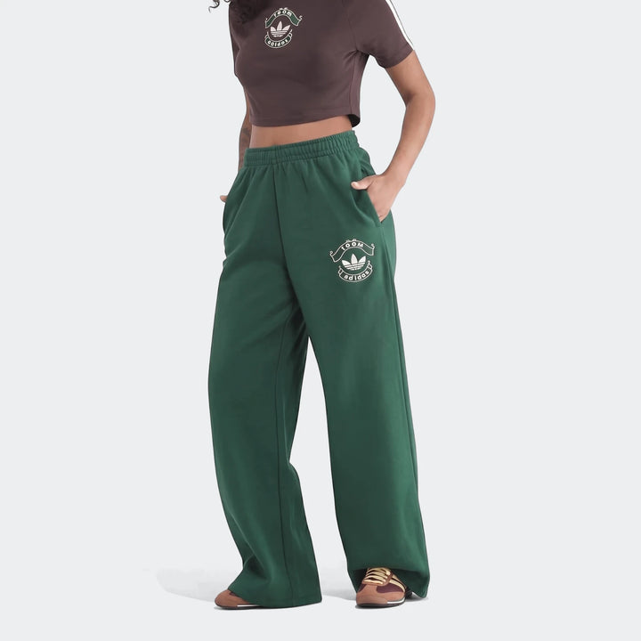 A model wears loose-fit green Infill Heritage Pants and a brown cropped t-shirt, standing with hands in pockets.