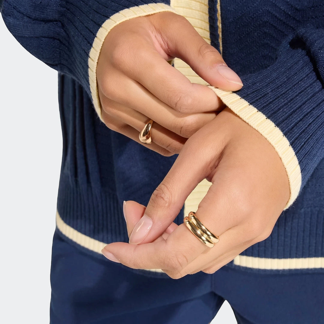 Close-up of hands wearing gold rings and a navy blue cable knit cardigan with cream trim, highlighting the intricate knit
