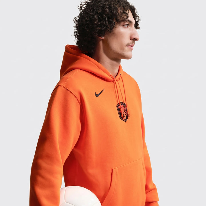 Male model wearing an orange Nike Netherlands Club Men's Soccer Pullover Hoodie with a white soccer ball.