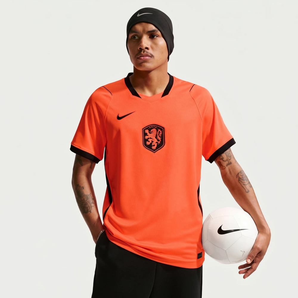 Male model wearing a bright orange Netherlands National Team 2026 Stadium Home Nike Dri-Fit soccer jersey, holding a white