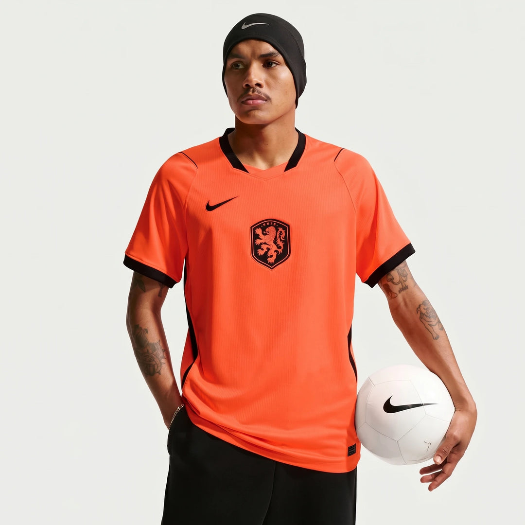 Male model wearing a bright orange Netherlands National Team 2026 Stadium Home Nike Dri-Fit soccer jersey, holding a white