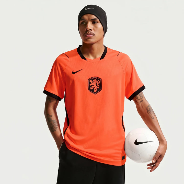 Male model wearing a bright orange Netherlands National Team 2026 Stadium Home Nike Dri-Fit soccer jersey, holding a white