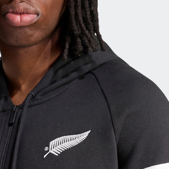 Close-up of a person wearing a black New Zealand Rugby Leisurewear Full Zip Hoodie with an embroidered silver fern logo.