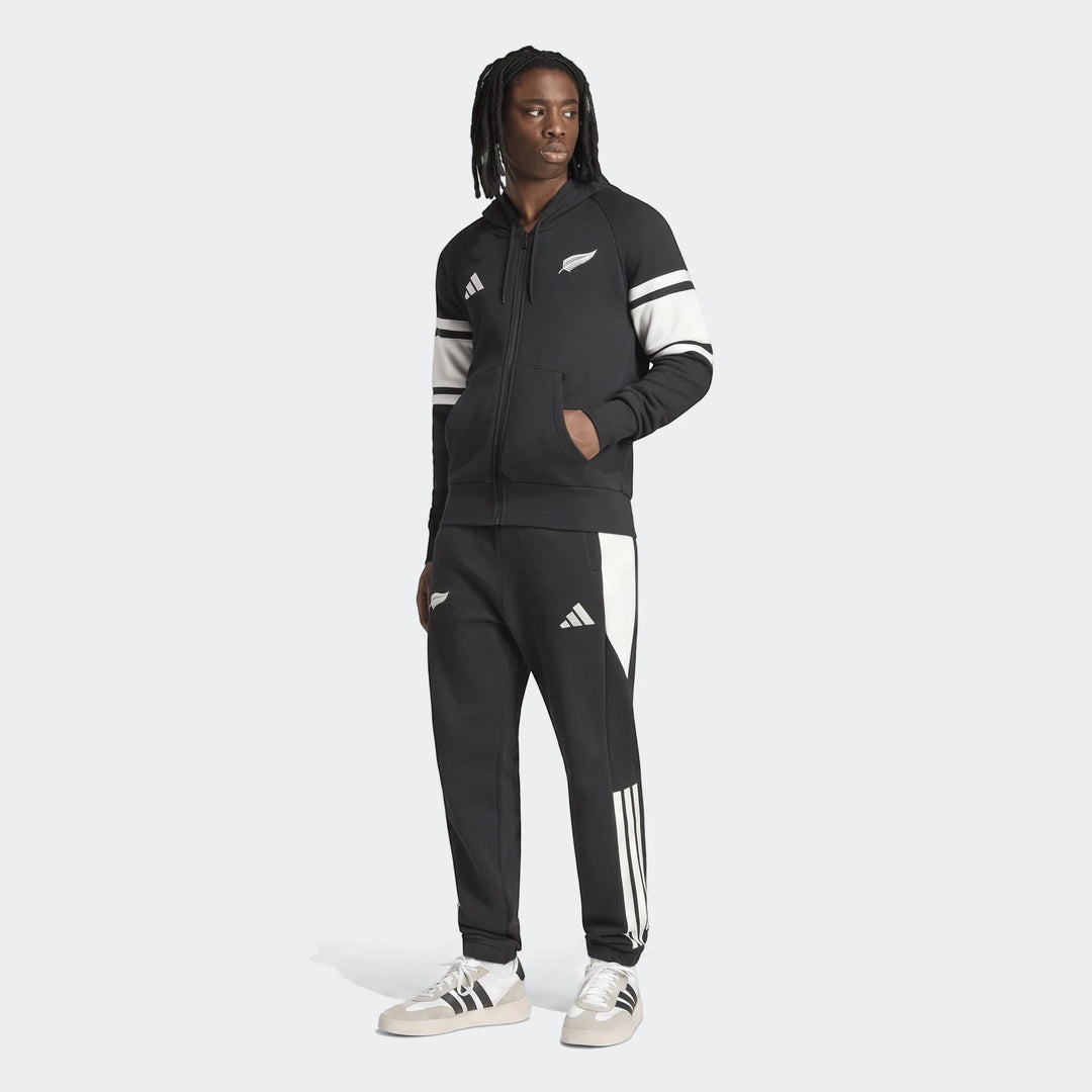 Man wearing a black New Zealand Rugby Leisurewear Full Zip Hoodie with white stripes, paired with black track pants and