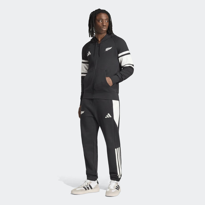 Man wearing a black New Zealand Rugby Leisurewear Full Zip Hoodie with white stripes, paired with black track pants and