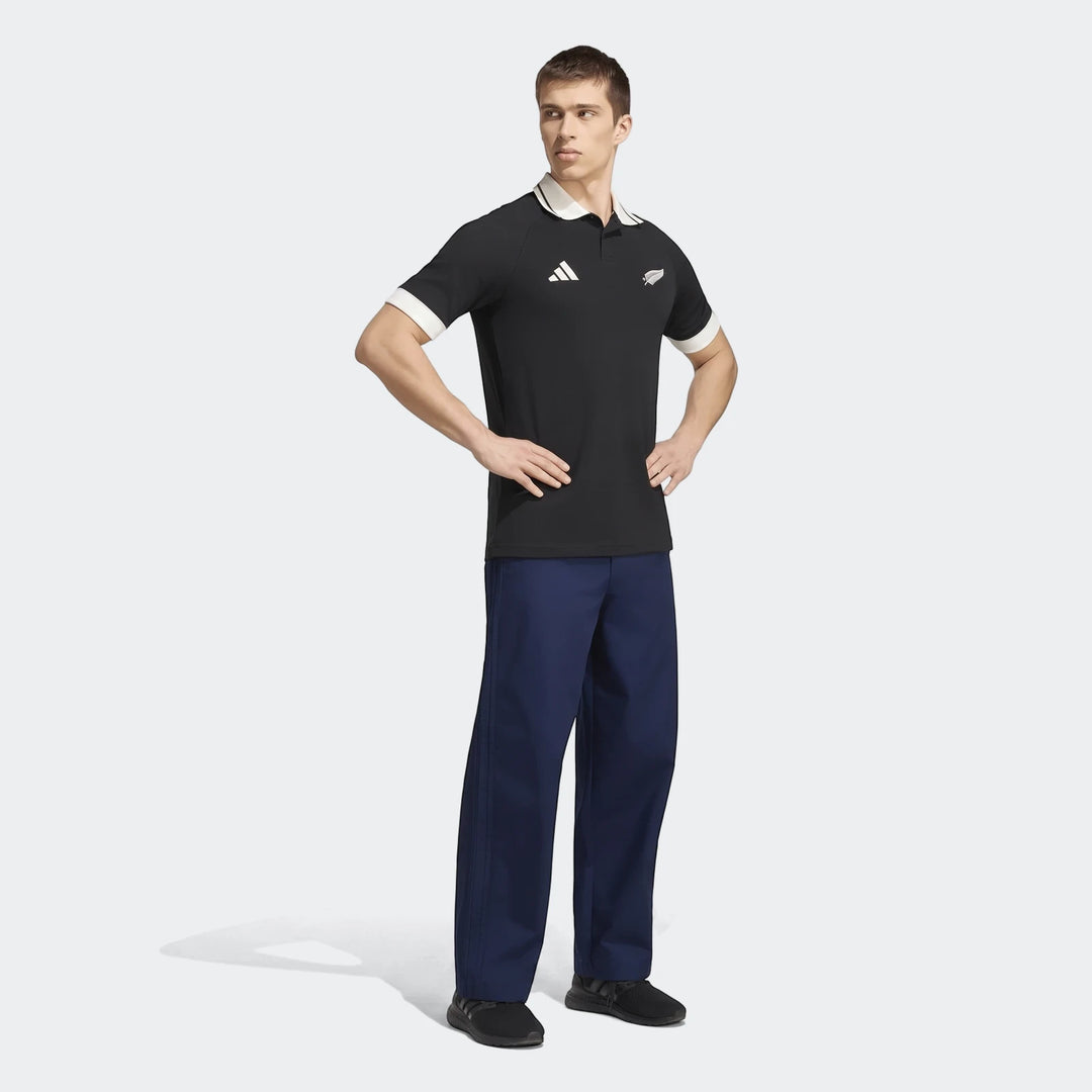 Male model in a black New Zealand Rugby Leisurewear Polo Shirt with white trim, dark blue pants, and black shoes, full body.