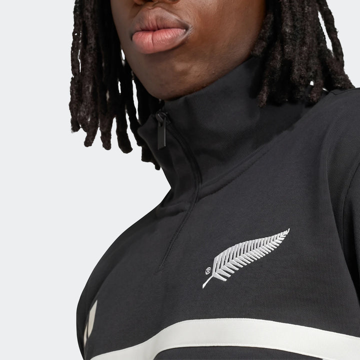 Close-up of a black New Zealand Rugby Leisurewear Quarter Zip Sweatshirt with a silver fern logo and white stripe, worn by a