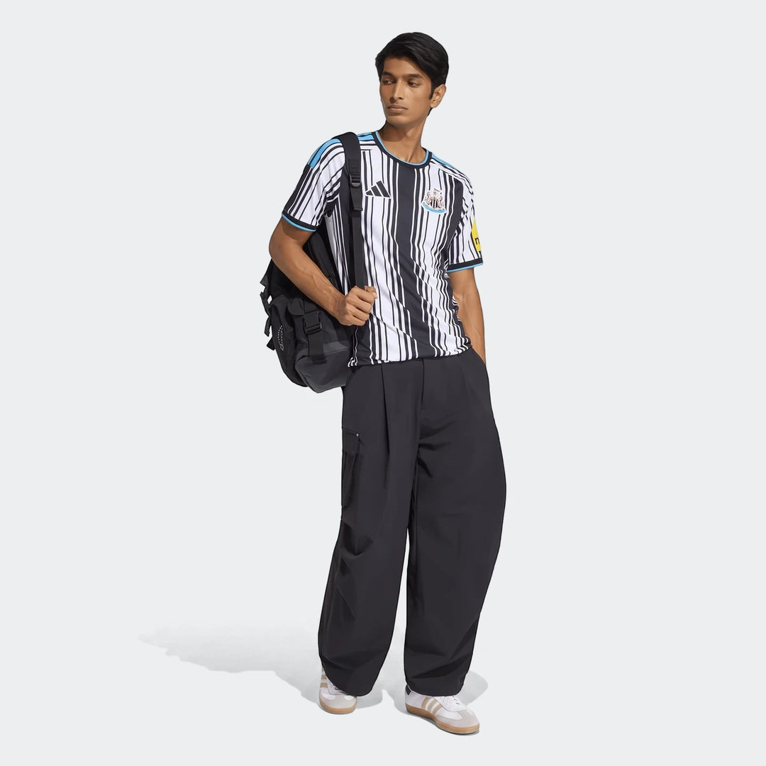 Male model wearing a Newcastle United Fc 26/27 Home Jersey, black cargo pants, and a black backpack, full body shot.