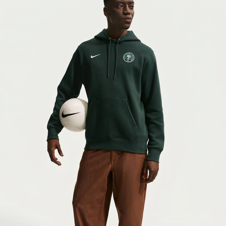 A man wears a dark green Nike Nigeria Club soccer pullover hoodie, featuring the Nike swoosh and NFF logo, holding a white