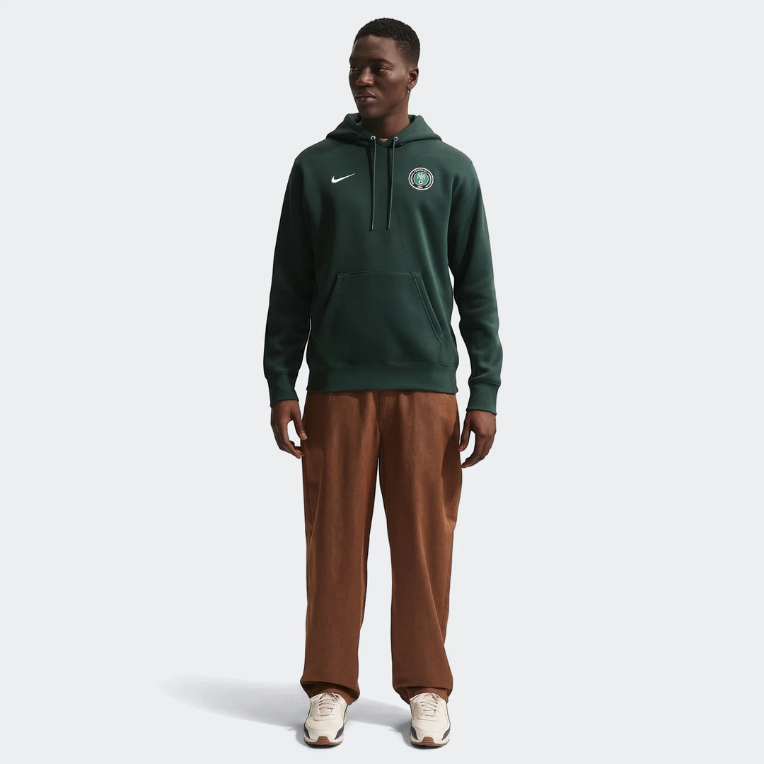 Dark green Nigeria Club Nike men's soccer pullover hoodie on a male model, styled with brown pants and sneakers.