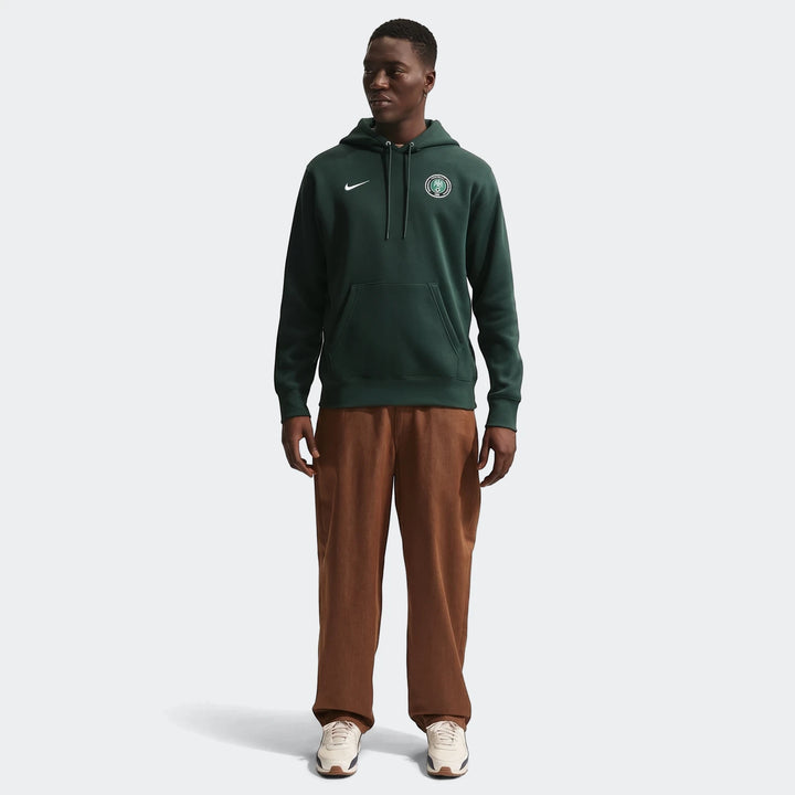 Dark green Nigeria Club Nike men's soccer pullover hoodie on a male model, styled with brown pants and sneakers.