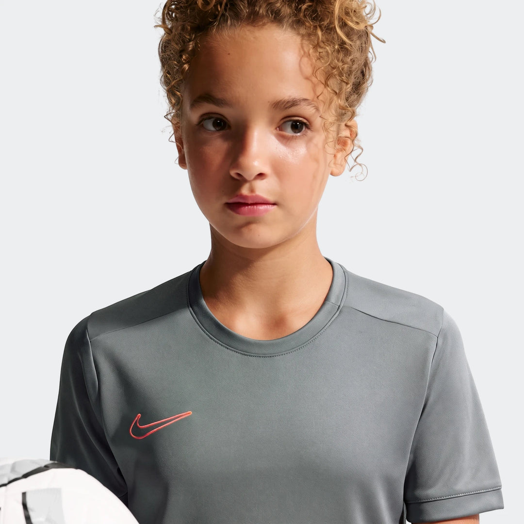 Young person in a grey Nike Academy Big Kids' Dri-Fit Soccer Top, short-sleeve, with a red Nike swoosh logo, holding a