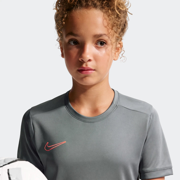 Young person in a grey Nike Academy Big Kids' Dri-Fit Soccer Top, short-sleeve, with a red Nike swoosh logo, holding a