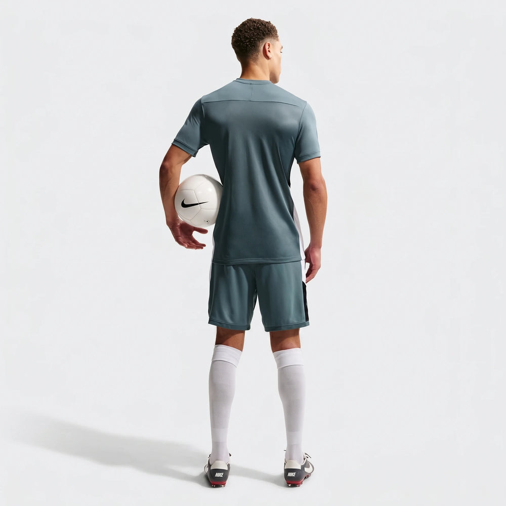 A man wears a green Nike Academy Dri-Fit short-sleeve soccer top, matching shorts, white socks, and cleats, holding a white