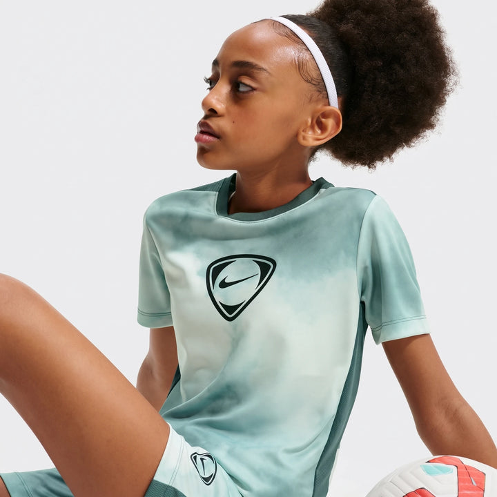 Young girl wearing Nike Academy+ Green Big Kids' Dri-Fit Short-Sleeve Soccer Top and matching shorts, sitting with a soccer