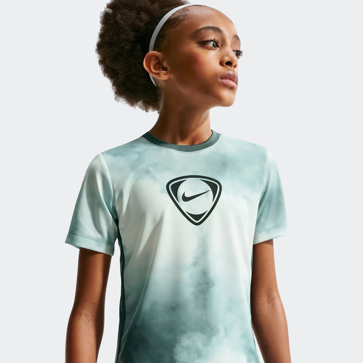 Young girl models Nike Academy+ Green Big Kids' Dri-Fit Short-Sleeve Soccer Top, featuring a tie-dye pattern and Nike logo.