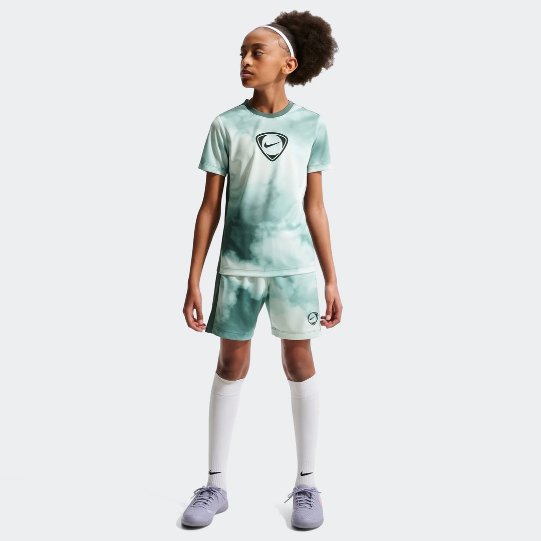 Young girl models Nike Academy+ green and white tie-dye Dri-Fit short-sleeve soccer top, matching shorts, and purple cleats.