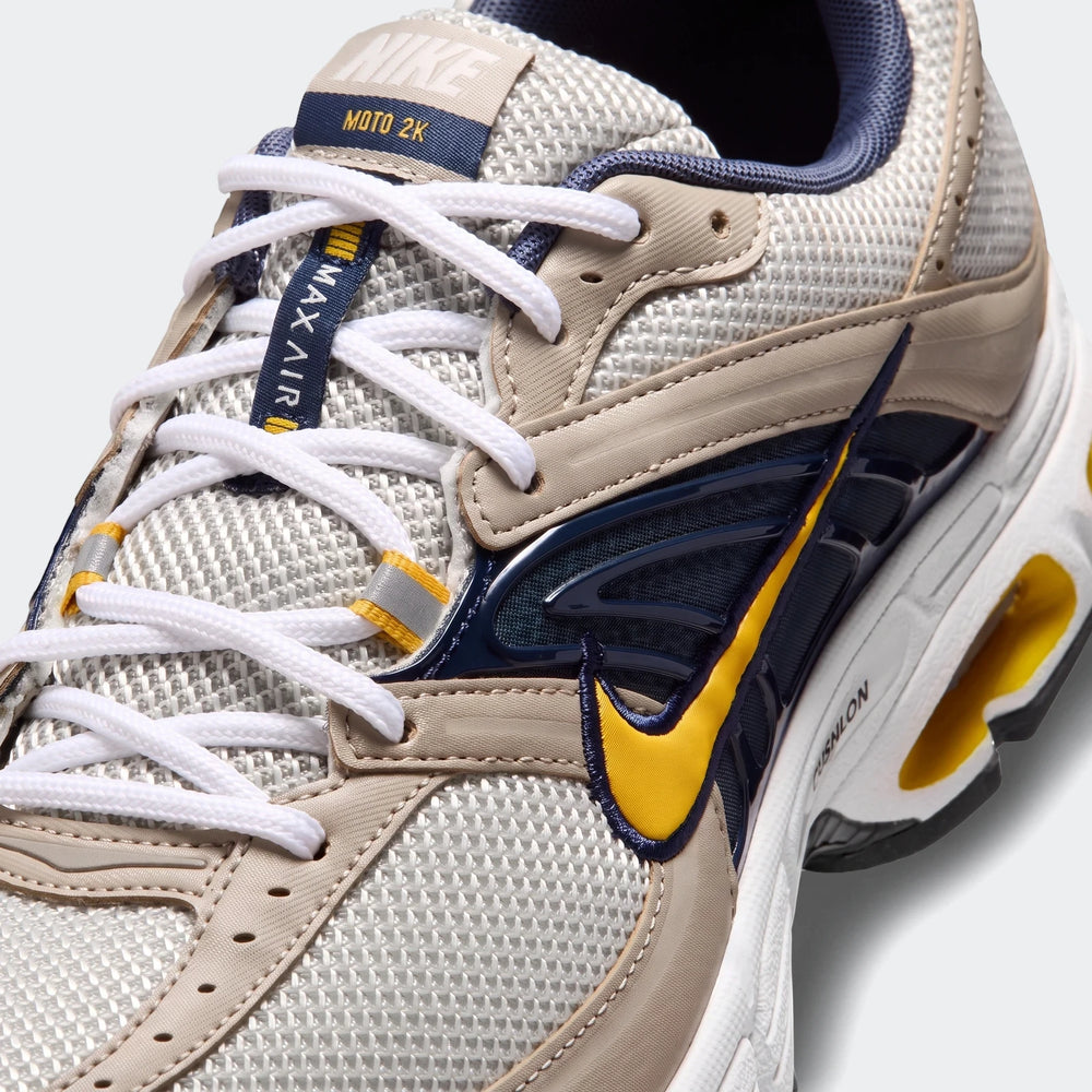 Close-up of a Nike Air Max Moto 2K 2Tone Men's Shoe in beige, white, navy, and yellow, showcasing the mesh upper and swoosh.