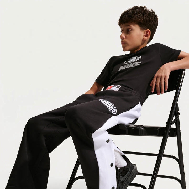 A young boy models black and white Nike Big Kids' Basketball Pants with snap buttons down the side, sitting on a chair.