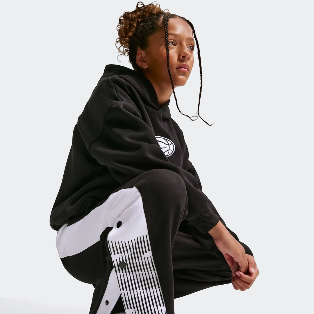 Young person in Nike Big Kids' black and white basketball pants and a black hoodie, shown in a crouching pose.