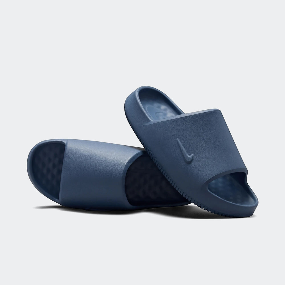 Two dark blue textured Nike Calm 2.0 Men's Slides, one upright with a swoosh logo and one on its side, full side view.
