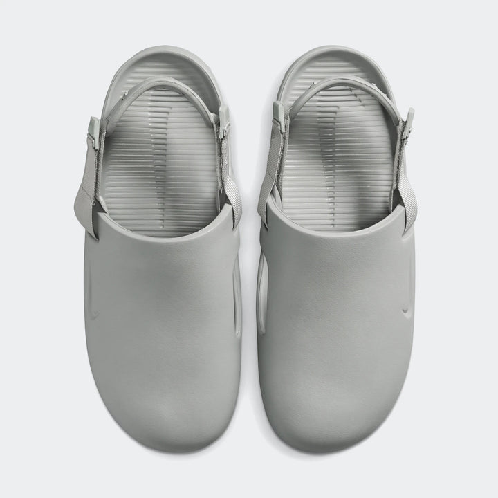A pair of light grey Nike Calm men's mules, featuring adjustable straps and a ribbed footbed, presented in a top-down view.