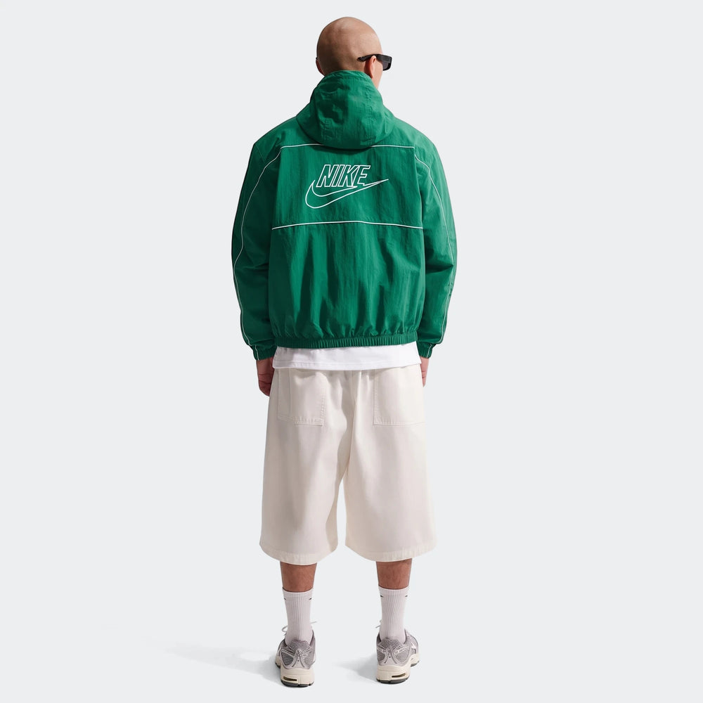 Back view of a man wearing a Nike Club green hooded jacket, paired with white shorts and sneakers on a plain background.