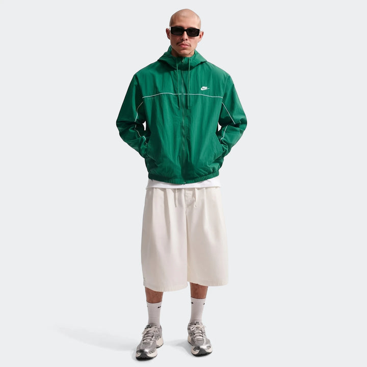 Full-body shot of a man wearing a green Nike Club hooded jacket, styled with off-white shorts and silver sneakers.