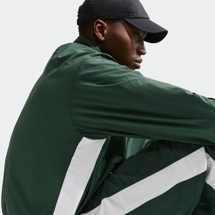 Dark-skinned male model wearing a dark green Nike Club Men's 1/2 Zip Jacket with white stripes and a black cap in profile.