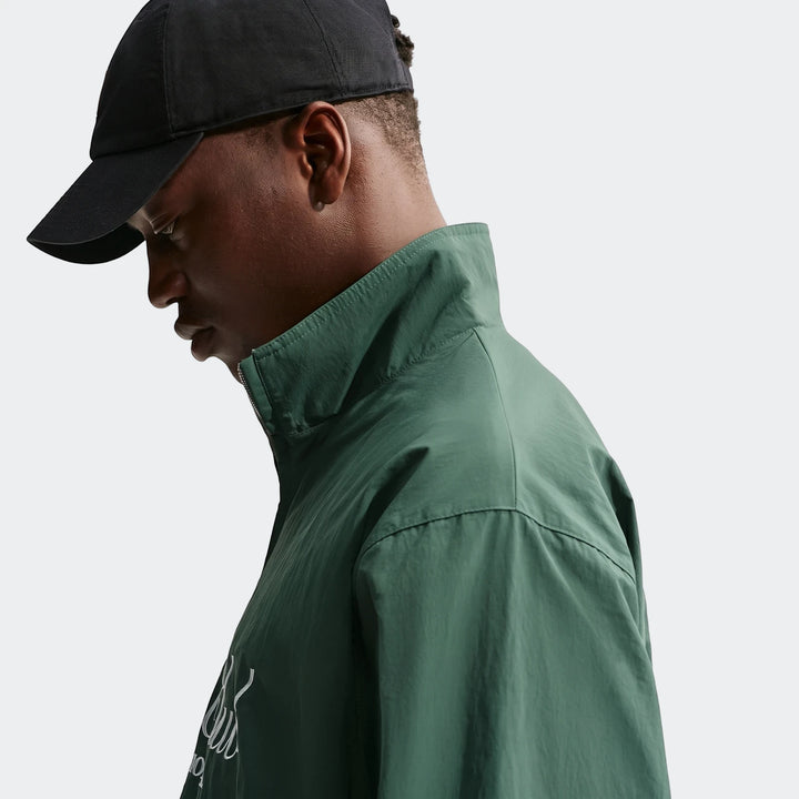 Nike Club Men's 1/2 Zip Jacket in green, worn by a man with a black cap, shown in a side profile view.