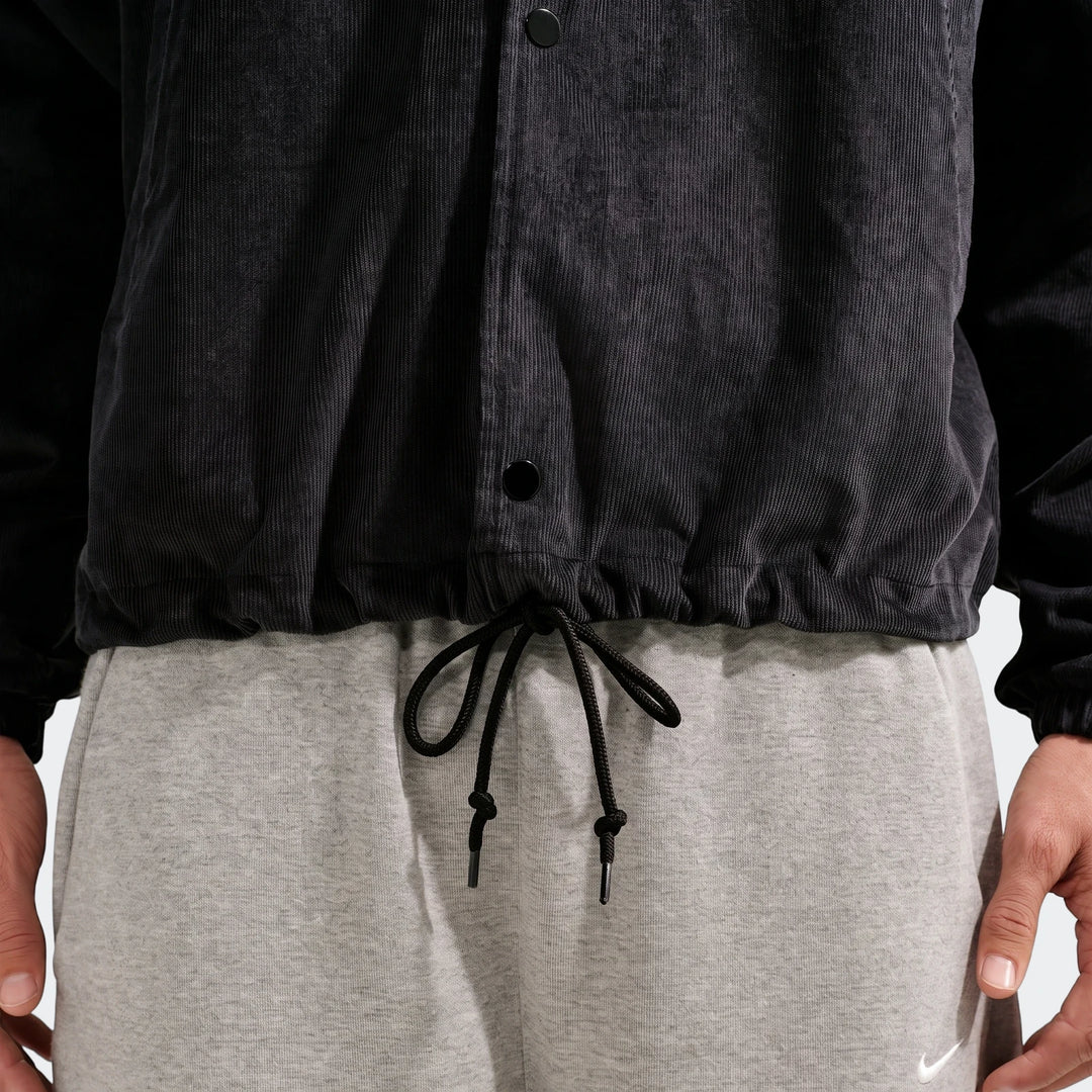 Close-up of a person wearing a Nike Club men's black corduroy coaches jacket with grey sweatpants and a white Nike swoosh.