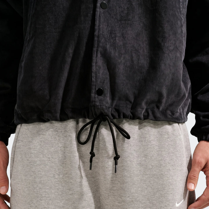 Close-up of a person wearing a Nike Club men's black corduroy coaches jacket with grey sweatpants and a white Nike swoosh.