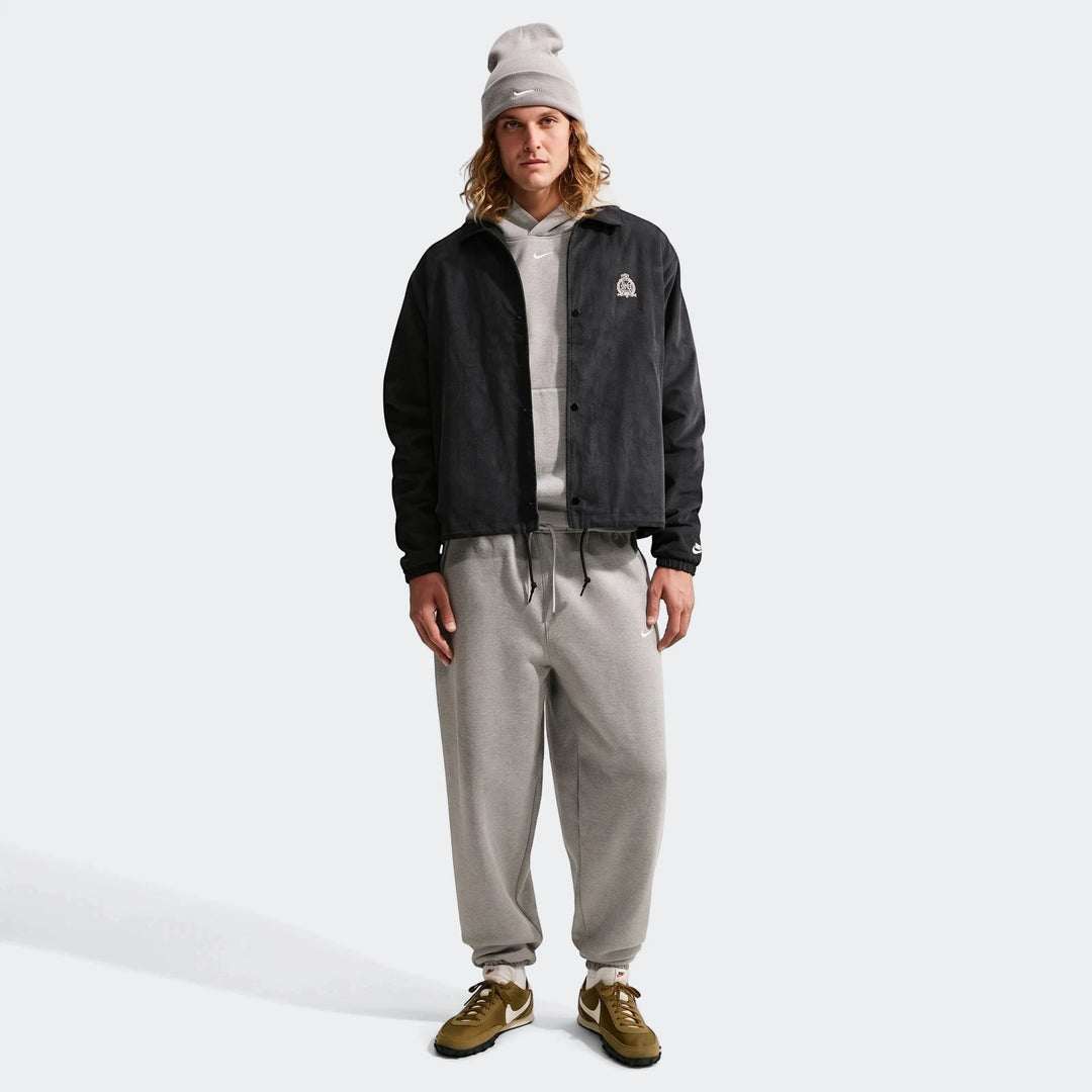 A man models the black Nike Club men's corduroy coaches jacket, styled with a grey hoodie, grey sweatpants, and olive green