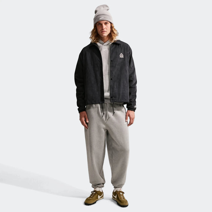 A man models the black Nike Club men's corduroy coaches jacket, styled with a grey hoodie, grey sweatpants, and olive green