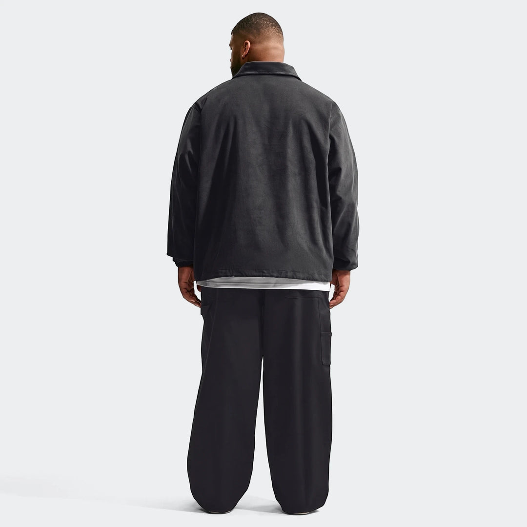 Back view of a man wearing a black Nike Club Men's Corduroy Coaches Jacket and black wide-leg cargo pants on a white